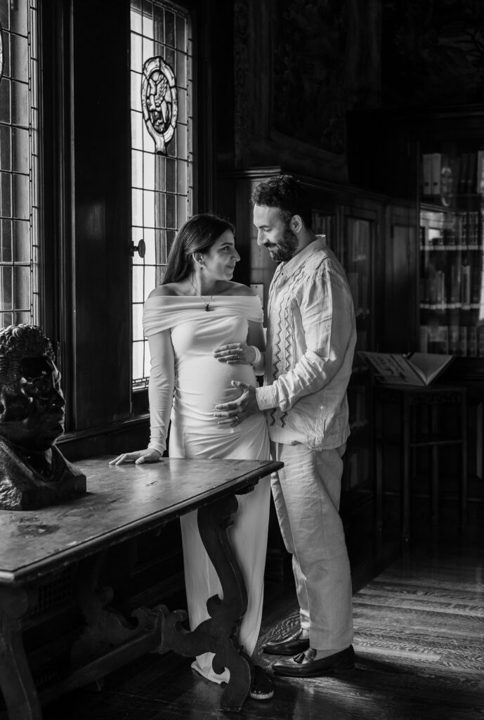 Husband gently holding his wife's baby bump as they stand together in the library of the Mexican Cultural Institute.